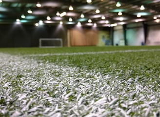 An indoor sports facility with artificial turf and overhead lighting. The focus is on the white boundary line on the field, and a soccer goal is visible in the distance. The lighting creates a well-illuminated environment with shadows cast on the turf.