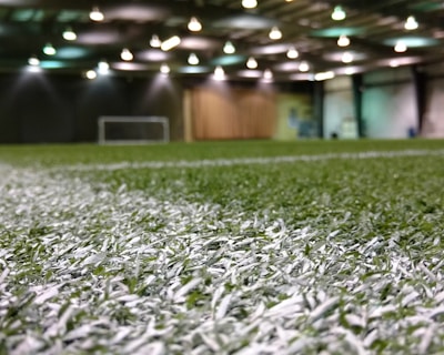 An indoor sports facility with artificial turf and overhead lighting. The focus is on the white boundary line on the field, and a soccer goal is visible in the distance. The lighting creates a well-illuminated environment with shadows cast on the turf.