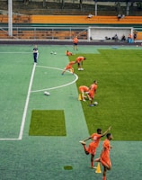 A group of people wearing bright orange sportswear are practicing on a green field. They appear to be athletes, likely soccer players, stretching and preparing for training. There are several soccer balls on the field, and a coach is observing from a distance. The field is surrounded by bleachers with a few onlookers seated, and the area is bordered by trees and residential buildings in the background.