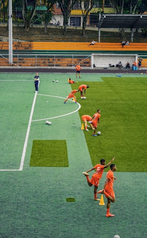 A group of people wearing bright orange sportswear are practicing on a green field. They appear to be athletes, likely soccer players, stretching and preparing for training. There are several soccer balls on the field, and a coach is observing from a distance. The field is surrounded by bleachers with a few onlookers seated, and the area is bordered by trees and residential buildings in the background.