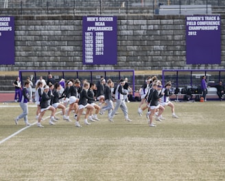 A group of athletes in sports attire are exercising or warming up on a grassy field. They are in a formation, appearing to be mid-stride, and the background shows a series of purple banners with sports achievements listed. The area is surrounded by a stone wall with benches and equipment visible.
