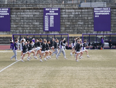 A group of athletes in sports attire are exercising or warming up on a grassy field. They are in a formation, appearing to be mid-stride, and the background shows a series of purple banners with sports achievements listed. The area is surrounded by a stone wall with benches and equipment visible.