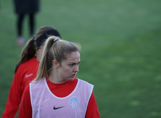 A person with a ponytail wearing a red sports jacket and a white training bib stands on a grassy field. The individual appears to be focused or concentrating, with others in similar attire visible in the background.