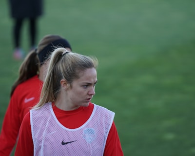 A person with a ponytail wearing a red sports jacket and a white training bib stands on a grassy field. The individual appears to be focused or concentrating, with others in similar attire visible in the background.