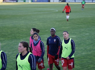 A group of female soccer players wearing training vests and sports attire walking on a green soccer field. Some players are wearing pink vests while others are in neon green. They are participating in a training session, with one player slightly in the background. The field is large and appears well-maintained, bordered in the distance by advertising boards.