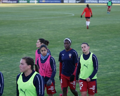 A group of female soccer players wearing training vests and sports attire walking on a green soccer field. Some players are wearing pink vests while others are in neon green. They are participating in a training session, with one player slightly in the background. The field is large and appears well-maintained, bordered in the distance by advertising boards.