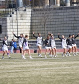 A group of athletes warming up or practicing on a grassy field near a track. They are wearing athletic gear, including shirts and shorts, and appear to be involved in a group exercise or routine. Most have one arm raised, and they are positioned in a line with a large stone wall in the background. A sign on the wall reads 'CAA', indicating a sports conference or event.