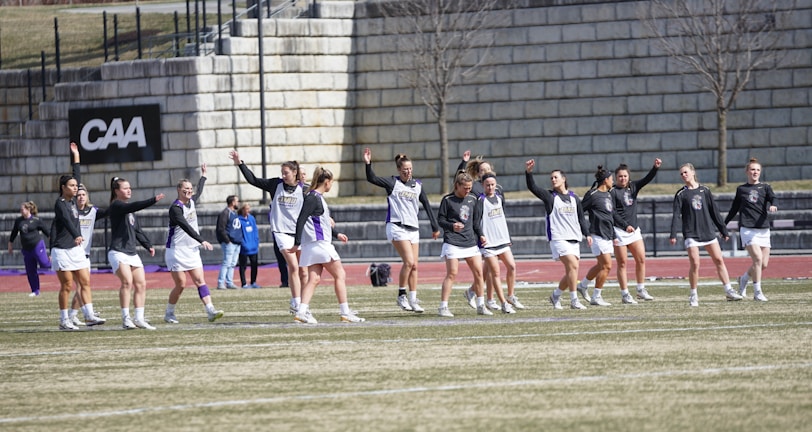 A group of athletes warming up or practicing on a grassy field near a track. They are wearing athletic gear, including shirts and shorts, and appear to be involved in a group exercise or routine. Most have one arm raised, and they are positioned in a line with a large stone wall in the background. A sign on the wall reads 'CAA', indicating a sports conference or event.