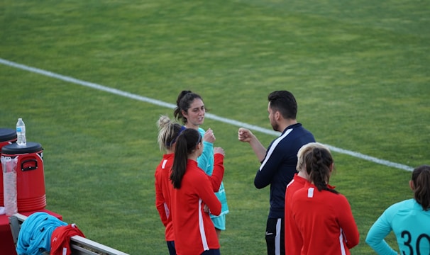 A group of people wearing sports attire are gathered on a grassy field. Some individuals are dressed in red jackets, while others are in turquoise and navy blue. A coach appears to be giving instructions. Sports equipment, such as water bottles and a bench with towels, can be seen nearby.