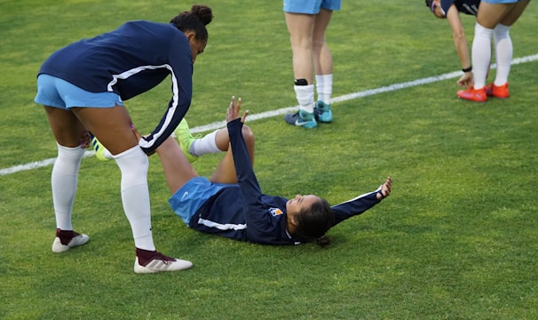 A group of athletes in sportswear are on a green soccer field. Two individuals are interacting closely; one helps the other who is lying on the grass. Nearby, other athletes are stretching.