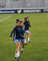 Three athletes are warming up on a grassy field, each wearing navy blue jackets and light blue shorts. They are engaged in a drill involving small orange and blue cones placed on the field. Spectators can be seen sitting in the background in a stadium setting, and advertisements are displayed on the side of the field.