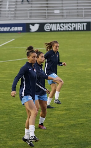 Three athletes are warming up on a grassy field, each wearing navy blue jackets and light blue shorts. They are engaged in a drill involving small orange and blue cones placed on the field. Spectators can be seen sitting in the background in a stadium setting, and advertisements are displayed on the side of the field.