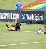 Two football players are performing a training drill on a grassy field. One is leaning back while controlling a soccer ball with her hands, and the other is lying on her back with her legs and arms up, holding another soccer ball. Both are wearing black attire with yellow socks. In the background, banners display social media handles and web addresses, and a person in a blue shirt stands nearby, watching the scene.