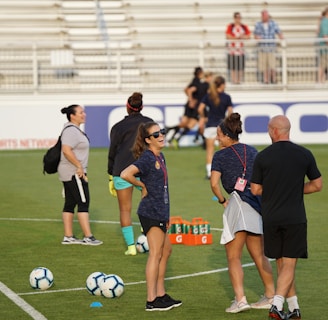 A group of people are gathered on a soccer field, likely during a training session. Some individuals are wearing athletic gear and appear to be coaching or instructing, while others are moving in the background. There are soccer balls scattered on the grass, and orange cones mark certain areas. A nearby bench is visible with some seated spectators.