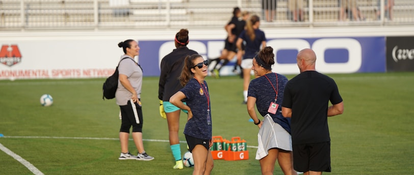 A group of people are gathered on a soccer field, likely during a training session. Some individuals are wearing athletic gear and appear to be coaching or instructing, while others are moving in the background. There are soccer balls scattered on the grass, and orange cones mark certain areas. A nearby bench is visible with some seated spectators.