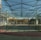 A spacious indoor sports facility with a tennis court and running tracks. The court is surrounded by high nets and tall, green metal structures supporting a roof. The area is well-lit with natural light filtering in through a transparent ceiling. In the background, there are trees with autumn foliage and several residential buildings.