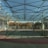 A spacious indoor sports facility with a tennis court and running tracks. The court is surrounded by high nets and tall, green metal structures supporting a roof. The area is well-lit with natural light filtering in through a transparent ceiling. In the background, there are trees with autumn foliage and several residential buildings.
