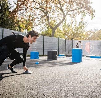 A man in athletic clothing is engaged in an outdoor workout or training session, bent forward while holding a marker or cone. The environment includes other sports equipment such as blue and gray barrels and more cones scattered around. In the background, additional people are seen participating, with trees and a building visible, suggesting a park or recreational area.