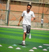 A person in sports clothing is running on a green artificial turf with white and red cones arranged in a pattern. The individual appears to be participating in a sports drill or exercise. The background includes a brick wall and netting consistent with an outdoor sports facility.