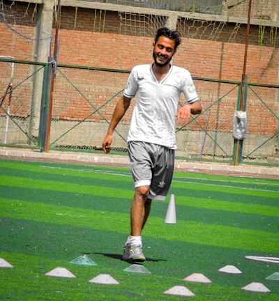 A person in sports clothing is running on a green artificial turf with white and red cones arranged in a pattern. The individual appears to be participating in a sports drill or exercise. The background includes a brick wall and netting consistent with an outdoor sports facility.