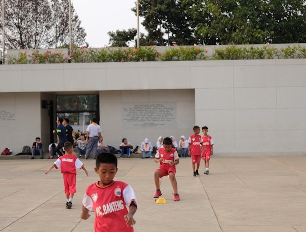 Several children in red sports uniforms are actively participating in a cone drill on a large concrete surface. The background features a modern white building, trees, and a few people sitting or standing near the structure.