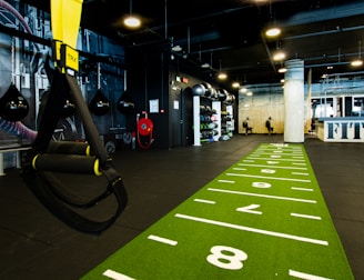 A modern gym space featuring a green track laid out on the floor with numbered lanes. TRX suspension trainers hang prominently in the foreground. The gym is equipped with various exercise equipment, including kettlebells and punching bags. In the background, there is a FIT sign near a reception desk under a low-ceilinged industrial design.