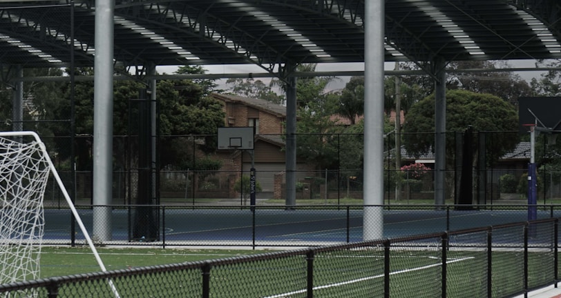 An outdoor sports facility featuring a running track and a soccer goal. The track is red, surrounded by a black metal fence, and the field is covered with artificial green turf. In the background, a basketball court is visible under a covered roof with metal supports. The sky is overcast, adding a moody atmosphere.