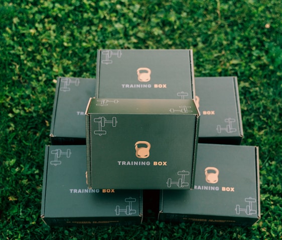 Several black boxes labeled 'Training Box' are stacked on a grassy surface. The top box features illustrations of a kettlebell and a dumbbell.