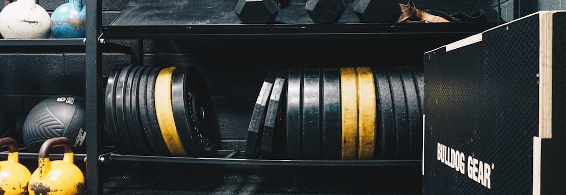 A gym setting featuring various fitness equipment, including kettlebells, stacked weight plates on a rack, dumbbells, and a medicine ball. The gym has a rugged, industrial vibe with black walls and a Bulldog Gear plyo box.