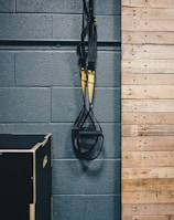 A pair of black and yellow suspension training straps hang from a sturdy metal support against a dark gray textured wall. A wooden panelled section is adjacent, with a black exercise box positioned on the floor.