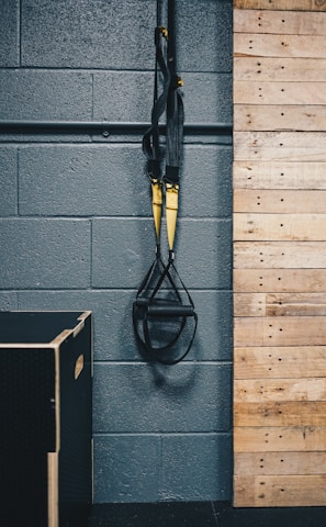 A pair of black and yellow suspension training straps hang from a sturdy metal support against a dark gray textured wall. A wooden panelled section is adjacent, with a black exercise box positioned on the floor.